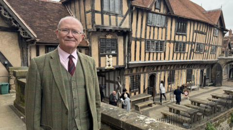 A man wearing a green suit stands in front of building. The building has a thatched roof and panels. Benches and fences can be seen to the right.