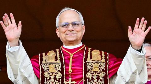 Pope Leo XIV, Robert Prevost wearing ornate ceremonial robes with gold embroidery stands with both arms raised. He is smiling and wearing glasses.