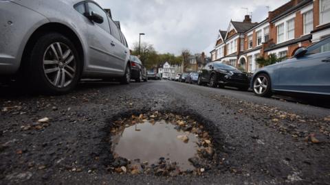 Pothole with stones in middle of residential street with cars parked either side