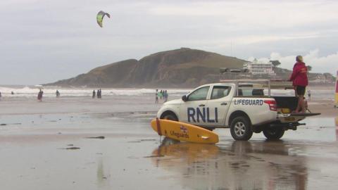 A beach scene featuring an RNLI lifeguard vehicle parked on wet sand with a yellow rescue board leaning against its front. A person in a red jacket stands near the back of the vehicle, facing the sea. In the background, beachgoers are visible in the water and on the sand, along with a kite surfer. Hills and buildings can be seen in the distance.
