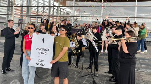 A band outside the Senedd with a poster saying 1 diwrnod i fynd, 1 day to go