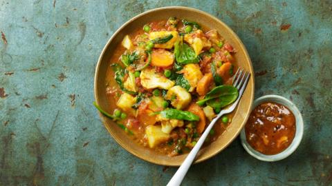 a brown bowl filled with vegetable curry with a fork, on a mottled green background. 
