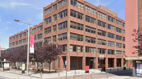 Vancouver House, a six storey red brick building with shutters on windows to the ground floor. It sits on a corner with pillars at the ground floor rising to the overhanging upper floors.