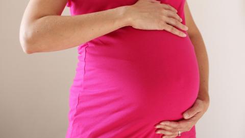 The torso of a woman wearing a bright pink dress who is eight months pregnant. She has her hands on the top and bottom of her baby bump.