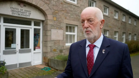 Steve Luce looking to the left wearing a navy blue blazer and striped shirt. He is wearing a red tie with white dots. In the background is a doorway with the words 'States of Jersey Department of Environment' above it. The building is light-brown brick with white frames around its windows, and there are planters in front of it with. 