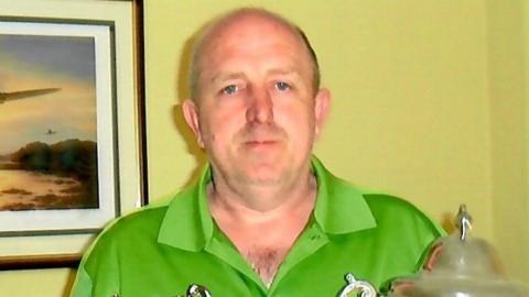 George in a green t-shirt and holding two trophies.