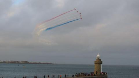 The Red Arrows flying over the lighthouse in St Peter Port. There are people standing on the pier and looking up near the lighthouse. 