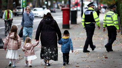 A family including two younger girls in white dresses and pink coats along with a little boy in a blue coat. They walk down the street with their backs to the camera and in the background two Metropolitan Police officers in uniform can be seen walking in the same direction.