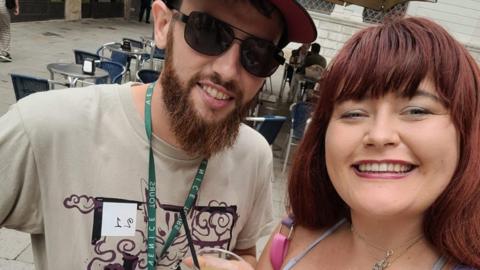 Chelsea Rees holding a drink. The mole is visible on her chest jus above her top. She is stood next to a man who has a beard and a beige t shirt on.