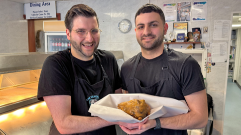 Two men with dark hair and beards in dark t-shirts holding a battered ball in white paper between them with silver counters and a white tiled wall behind them.
