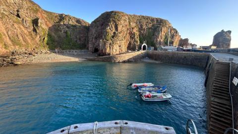Creux Harbour in Sark. Three small boats are moored up in the harbour. A yacht is berthed on the harbour wall.