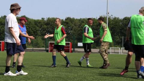 Veterans take part in a walking football session. They are on a pitch after the event, shaking hands and talking. Four of them are wearing green vests over their sports gear.