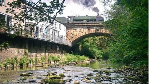 A railway bridge over the river with a steam train going across it. To the left of the picture are white buildings looking out to the river and bridge with a balcony. To the right of the picture are trees