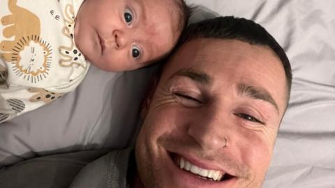 A baby in a sleepsuit with animals depicted lays on a bed with his smiling father looking up at the camera