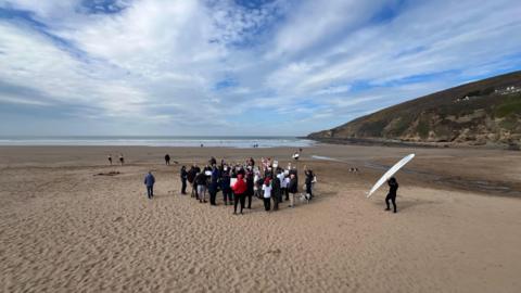 A picture of the protestors gathered on the beach raising their placards in the air. Behind them is a blue cloudy sky and a blue sea. A cliff can be seen to the right of the image.