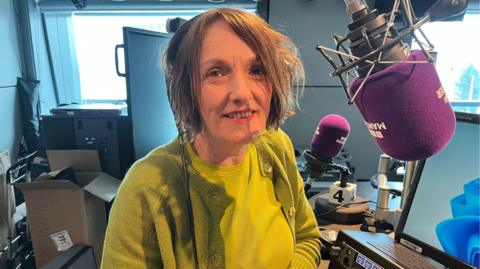 Jane Wilkinson, who is wearing a yellow top and green cardigan, is sitting in front of a BBC Radio Manchester purple branded microphone in the radio studio.