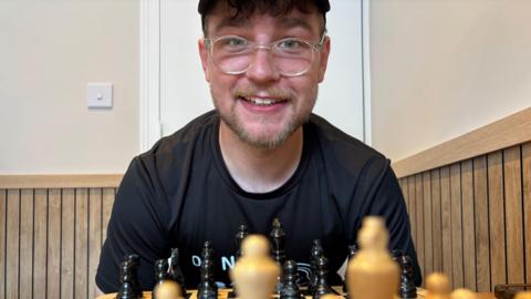A young bearded man with glasses smiles and leans down over a chess board.