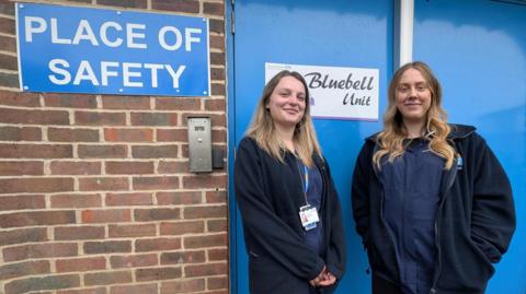 Two women in all-blue uniforms stand outside a blue door which says 'Place of Safety'. A brick wall is to the left of the image.