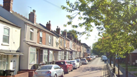 A street with terraced housing on the left and green trees on the right
