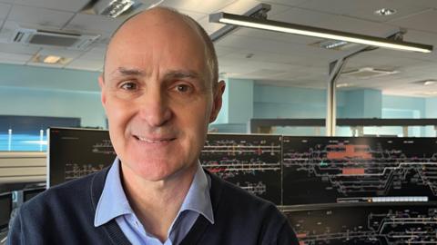 Kevin Wolloughby is on the left of the image. He is wearing a blue shirt with a navy jumper, and is standing in front of two large screens which are showing the railways in Cambridgeshire.