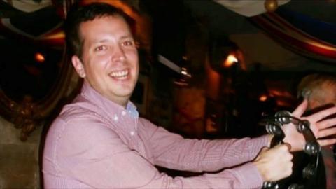 A man standing side on but looking at the camera. He is smiling and holding a tamborine with his arms out stretched. He appears to be in a dark venue with dim lighting.