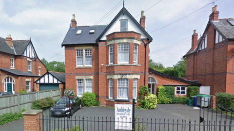 A screenshot from Google street view showing the care home. It is a large detached three-storey brick house with bay windows and a driveway outside the front. There are black railings around the property and a black car parked outside.