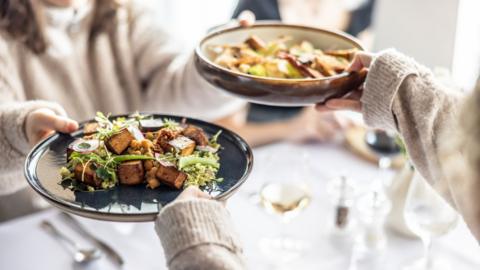 A moment of sharing a meal, where two people exchange dark bowls with various, richly decorated salad dishes.
