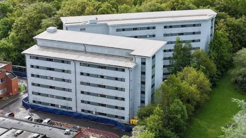 An exterior, aerial shot of Arrowe Park hospital - showing two large white multi-storey buildings surrounded by a car park and trees.