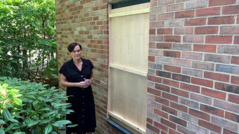 Sarah Sasmazer standing outside the broken office window. She is wearing a black dress and has dark short hair. The window has been boarded up with light-coloured wood. The window is at the side of a building and looks out at green bushes.
