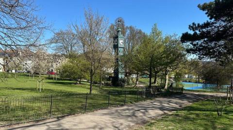 Blakers park in Brighton has a wrought‑iron fence and green lawn leading towards a tall ornamental clock tower, surrounded by trees, with a tennis court visible in the background under a clear blue sky.