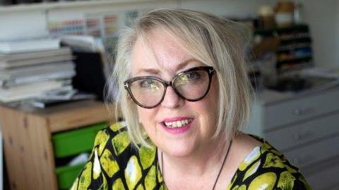 A woman with short blonde hair and large black-framed glasses. She is sitting down wearing a dress with yellow patterns. In the background there are cabinets with folders on top.