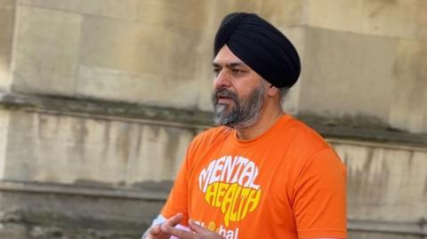 Bhupinder Sandhu looking to the left with a determined expression. He is wearing a black turban and an orange t-shirt that says "mental health" on it. Behind him is a sandstone wall. 
