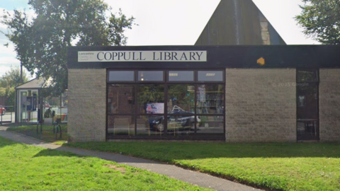 A street view of the exterior of Coppull Library. It is a grey-bricked building with dark framed windows. It has Coppull Library written across the top.