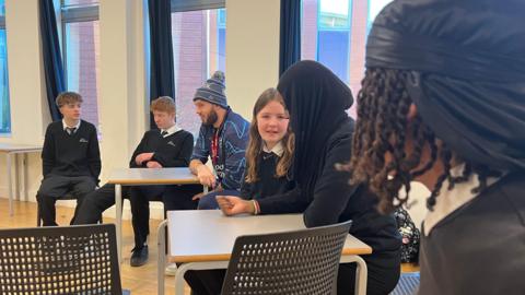 Six secondary school pupils sit around two small, square tables in a classroom alongside a man in Bristol Bears kit and beanie hat. The pupils are a mixture of boys and girls and wear dark school jumpers over white shirts and ties.