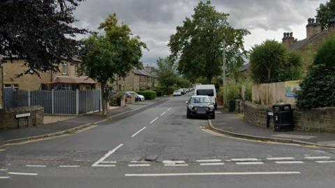 A road stretches into the distance, with cars parked on both sides. It is a residential street and homes with driveways and fences can be seen on both sides of the road. Trees line the street.
