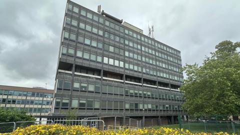 A grey seven-storey office block with lots of windows and metal frames with a tree and yellow flowers in the foreground against a cloudy sky