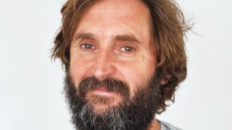 Comedian Joe Wilkinson. Dark brown facial hair and long brown hair. He is smiling at the camera