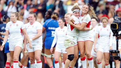 England's Megan Jones and Jess Breach celebrate after qualifying for the final.. They are wearing the white kit and hugging with other team members behind them and a camera to the right. Crowds are in the distance. 