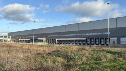 A shot of the new distribution centre - it is a large grey rectangular building with multiple lorry loading bays on the side and office buildings to the left