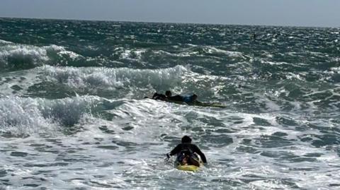 One of the surf lifeguards is on a paddleboard paddling out to two young people on surfboards in choppy water on a sunny day.