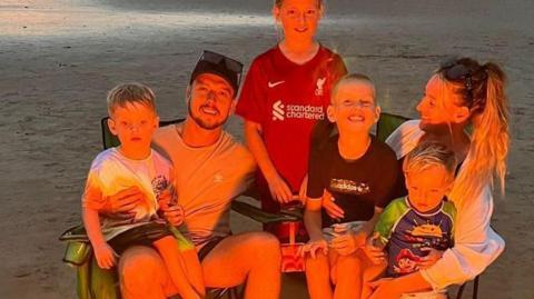 Nathan Osman and his partner Katie sitting on camping chairs with their four children at the beach. They are lit by the burning glow of the fire.