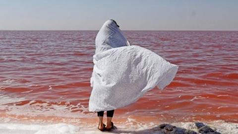 Person in a white shawl stands of the shoreline of Lake Urmia and its red-coloured water