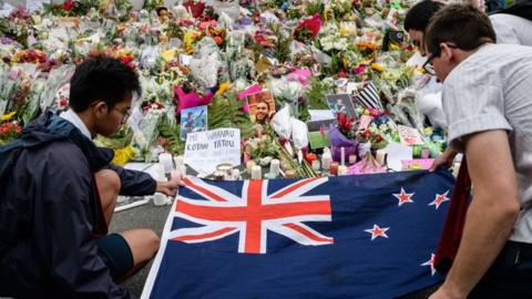 Mourners lay out a New Zealand flag next to a bed of floral tributes for Christchurch shooting victims in 2019