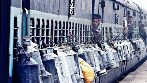 Milk cans are tied to the outside of a train as milkmen peep out of the doors.
