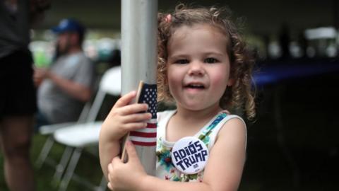 A two-year-old girl with a bade reading "Adiós Trump" stands at the Polk County Steak Fry