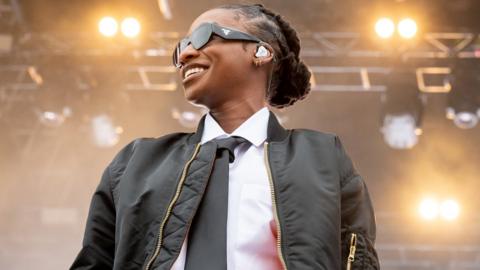 Little Simz performs at Parklife Festival 2023. Little Simz is a young black woman with braided hair tied back in a bun behind her head. She wears black Prada sunglasses, a black bomber jacket and black tie with a white shirt. She smiles as she looks to her right off stage. Behind her you can see stage lighting