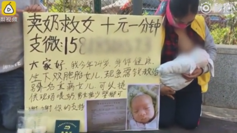 Woman kneels on street in China, breastfeeding a baby. The sign beside her has a picture of her ill baby and writing in Chinese characters. There are two plastic bags apparently filled with breast milk in the foreground.
