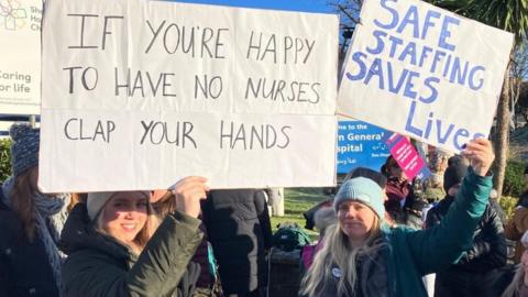 Nurses holding up signs on the picket line which say 'If you're happy to have no nurses clap your hands' and 'Safe staffing saves lives'