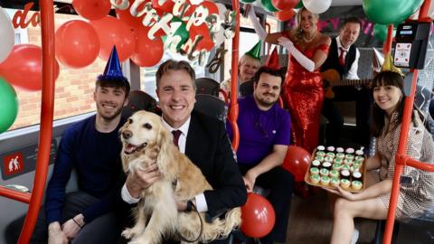 Metro mayor Dan Norris with a dog on a bus full of people and birthday balloons