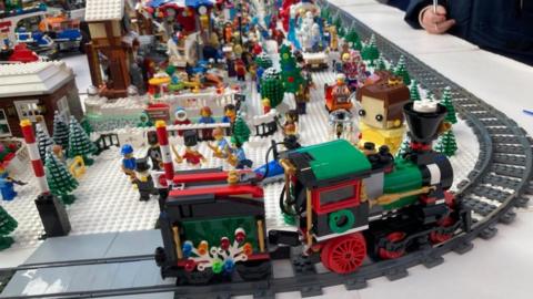 Lego train with a green and red engine moves along a railway track past groups of people